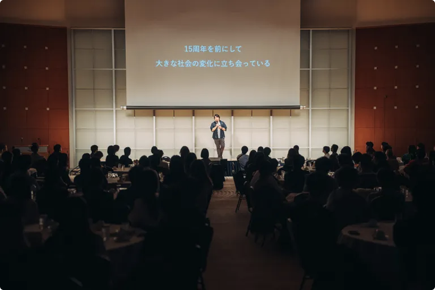会場で多くの観客が着席して登壇者の講演を聞いている様子。スクリーンには「15周年を前にして　大きな社会の変化に立ち会っている」と表示されている。