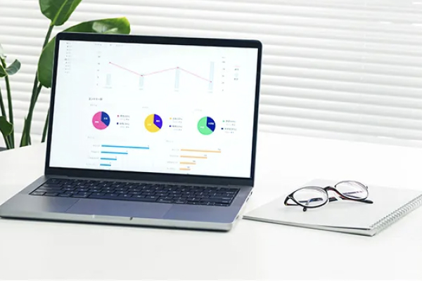 デスク上のノートパソコンにグラフやチャートが表示され、横にノートと眼鏡が置かれている様子。