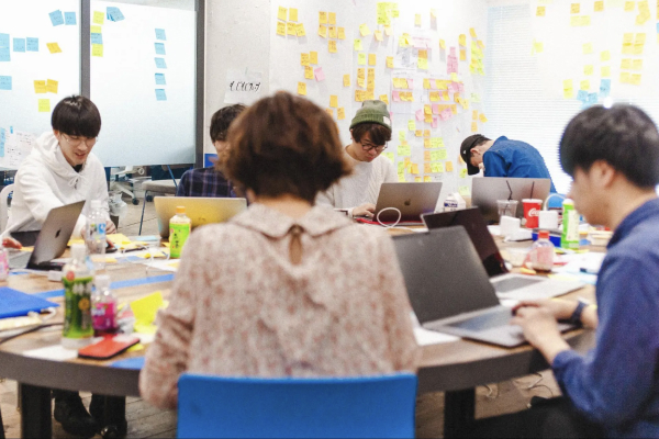 オフィス内でメンバーがノートパソコンを使い、付箋が貼られた壁を背にして作業や議論を行っている様子。