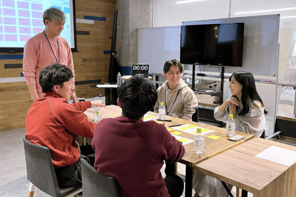 社内スペースで部門主導の勉強会が行われ、テーブルを囲んだメンバーが付箋や資料を使いながら意見交換している様子。