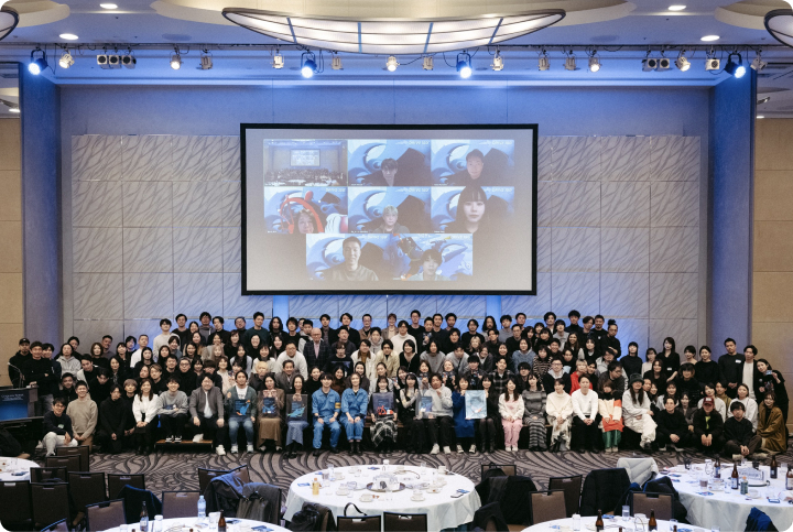 大人数のメンバーがステージ前に集まって撮影した集合写真。背後のスクリーンにはオンライン参加者の映像が映し出されている。