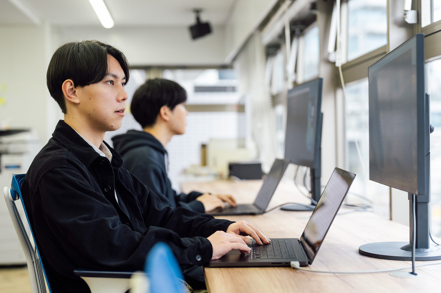 ノートPCと外付けモニターを前に、2名の社員がそれぞれ自席で作業している様子。