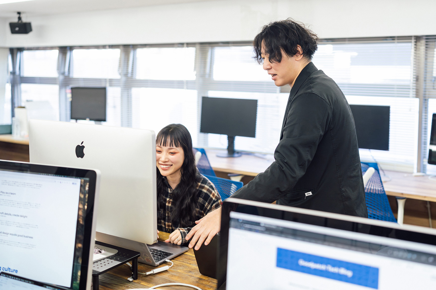 オフィスで、デスクに座る社員と、その横に立つ社員が同じモニター画面を見ながら作業している様子。