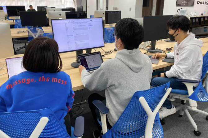 デスクに並んだ3人の社員が、ノートPCと外部モニターを見ながら同じ方向を向いて作業している様子。