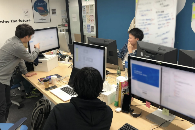 オフィスで1名の社員がモニターを指さし、他の2名の社員に説明している様子。