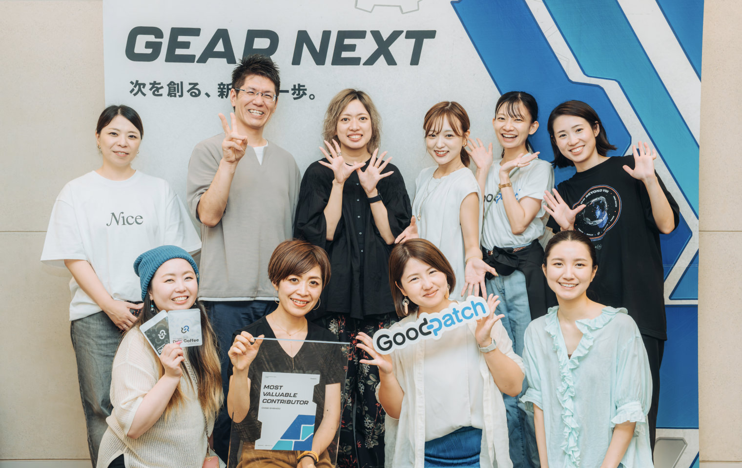 笑顔で集合写真を撮るメンバーたち。背景に『GEAR NEXT』のボードがあり、数名が手を挙げたり、ロゴ入りのボードを掲げたりしている
