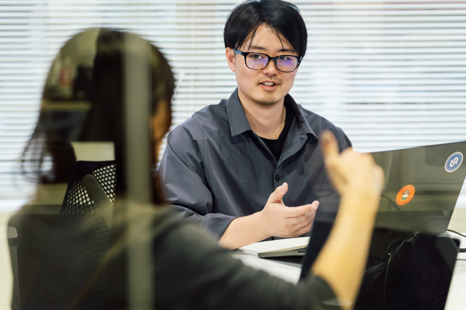 オフィスのデスクで、ノートPCを前に2人が向かい合って話している様子。