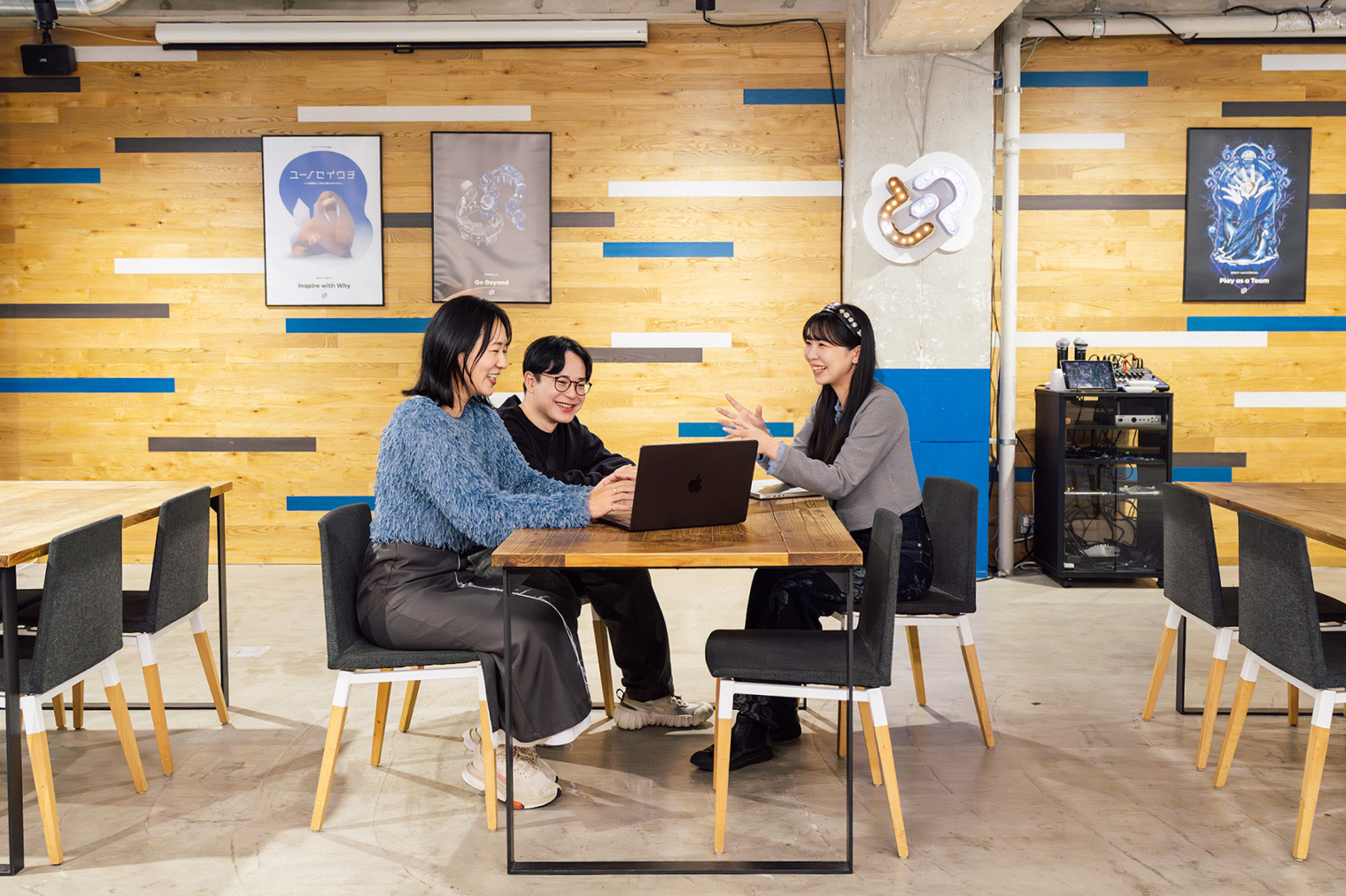 オフィス内で3人の社員がテーブルを囲み、ノートパソコンを見ながら打ち合わせをしている様子