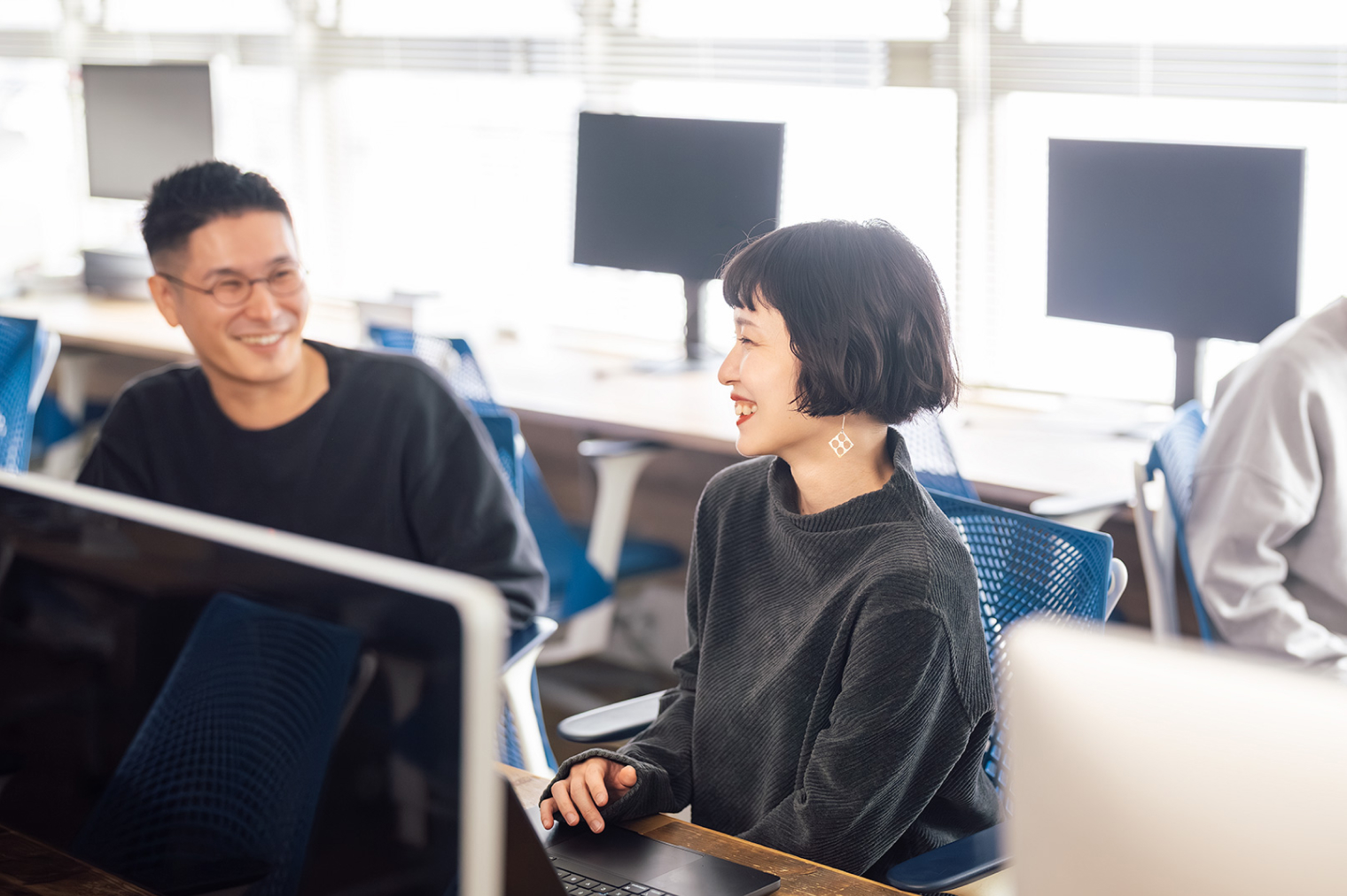 オフィス内で椅子に座り、会話をしながら笑顔でコミュニケーションを取る社員2名の様子。